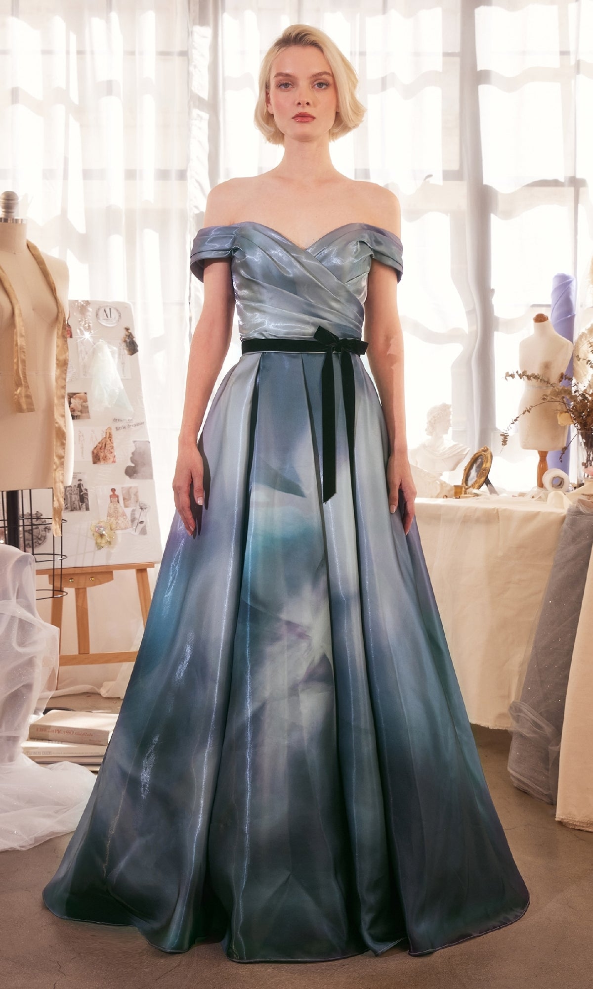 A woman stands indoors in the Off-Shoulder Blue Prom Gown: Andrea & Leo A1431, a floor-length blue dress with a black sash, surrounded by dress forms and sewing materials.